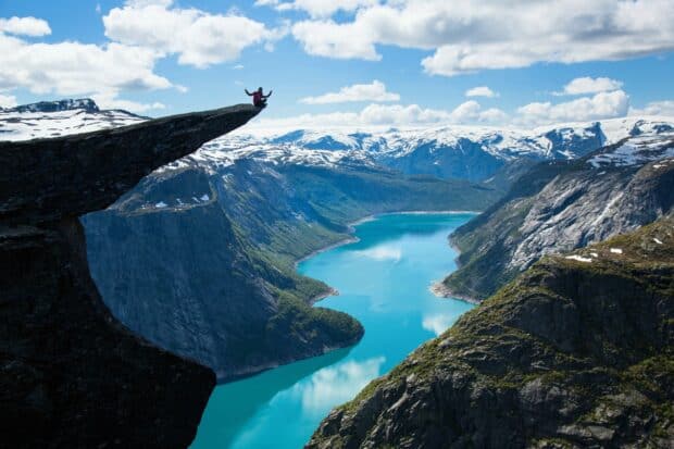 A person sits on a cliff overlooking the turquoise water and snowy mountains in Norwegian Fjords, HD Desktop Wallpaper