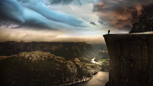 A person stands on a cliff overlooking Norwegian fjords with moody clouds above, 4K Desktop Wallpaper