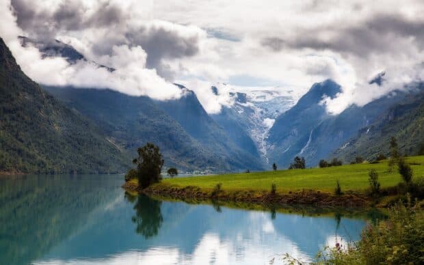 A calm water fjord surrounded by green fields and towering mountains under cloudy sky in Norway, HD Desktop Wallpaper