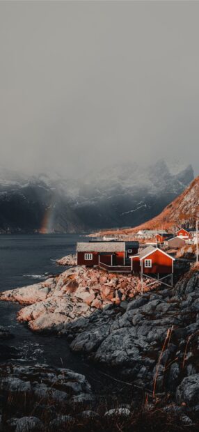 A scenic view of Norwegian fjords with red houses along the rocky shore and a faint rainbow under cloudy skies, HD Phone Wallpaper