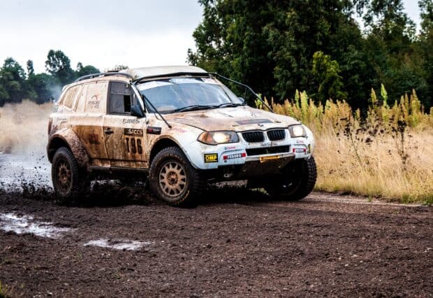 A rally raid car is driving fast on a dirt road surrounded by vegetation, 2K Desktop Wallpaper