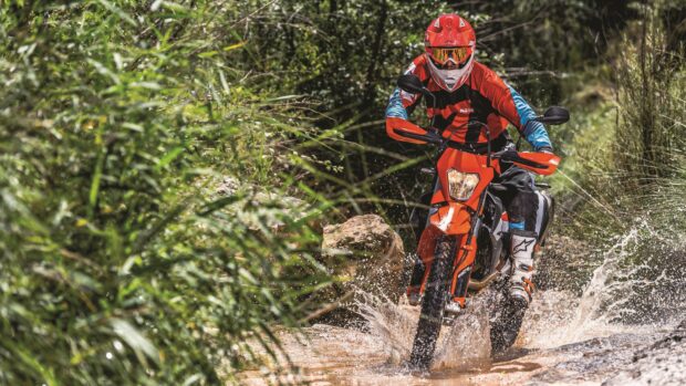 A rider on an Enduro motorbike splashes through a shallow water path surrounded by greenery, 4K Desktop Wallpaper