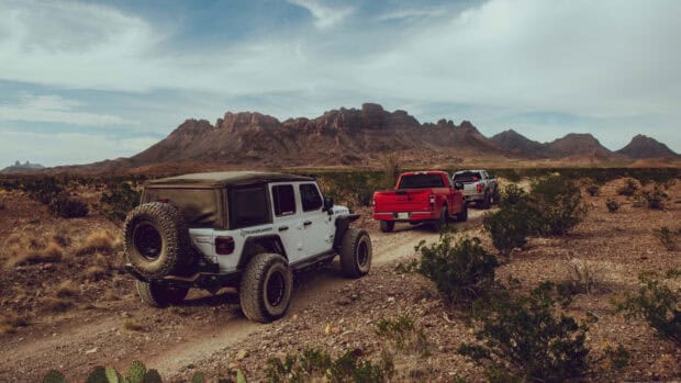 A convoy of off road vehicles is driving through a rocky desert trail near mountains under a cloudy sky, HD Desktop Wallpaper