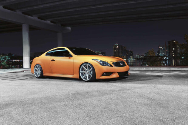 A sleek orange Infiniti G37 car is parked under a concrete structure with city lights in the background, HD Desktop Wallpaper