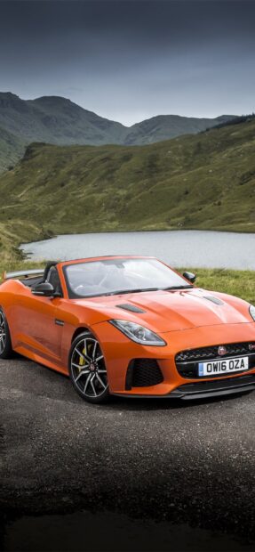 A stylish orange Jaguar F Type is parked near a lake with mountains in the background, 2K Phone Wallpaper