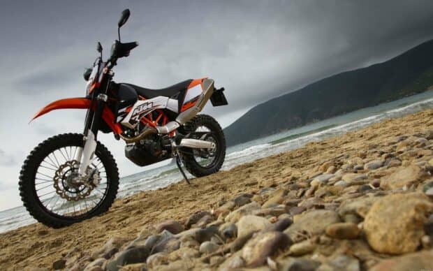 An orange KTM Enduro motorbike is parked on a sandy beach with rocky foreground and cloudy sky