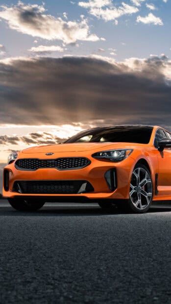 An orange Kia car is parked on a road under a dramatic cloudy sky with the sun setting, 4K Phone Wallpaper