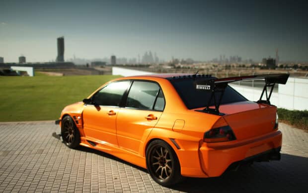 An orange Lancer Evo with a large rear wing parked near a green field in the city, HD Desktop Wallpaper