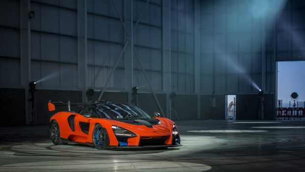 An orange McLaren Senna sports car is parked in a large industrial hangar with spotlights shining around it, 4K Desktop Wallpaper