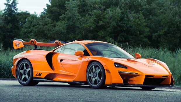 The orange McLaren Senna is parked on a paved road with green trees in the background, 2K Desktop Wallpaper