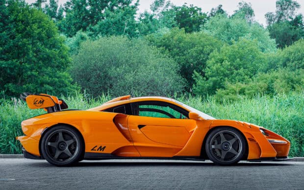 A bright orange Mclaren Senna is parked on a paved road near green trees, 2K Desktop Wallpaper