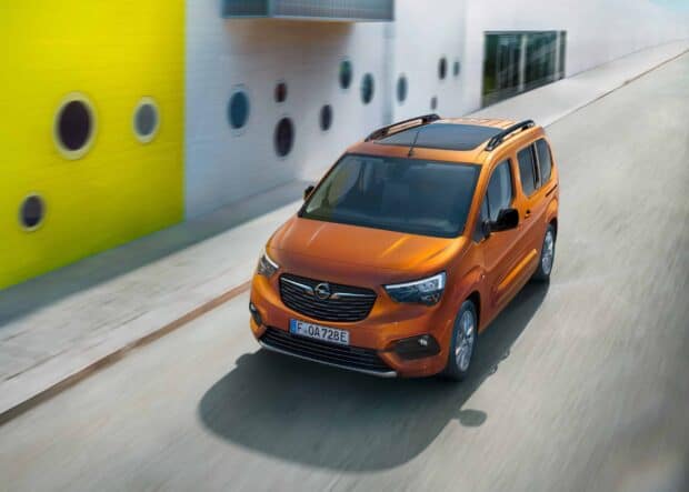 An orange Opel Combo driving smoothly near a building with circular windows and a yellow wall, 2K Desktop Wallpaper