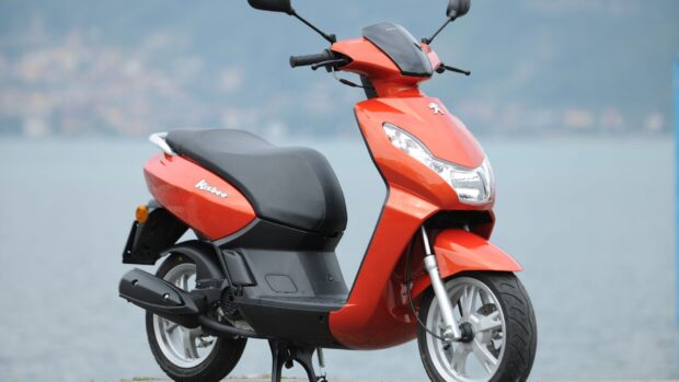 The orange Peugeot Kisbee scooter is parked beside a body of water with a blurred cityscape in the background, HD Desktop Wallpaper