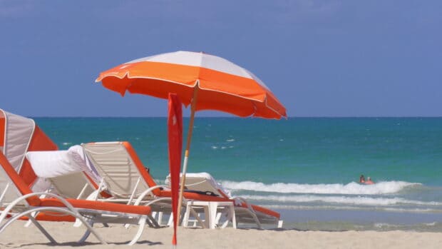 An orange beach umbrella stands beside lounge chairs by the blue ocean waves, 4K Desktop Wallpaper