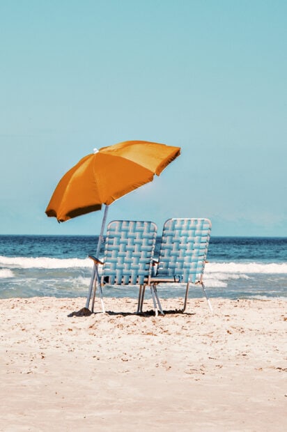 A bright orange beach umbrella shades two striped chairs on sandy shore by blue ocean, 2K Mobile Wallpaper