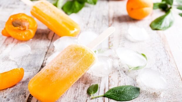 A bright orange popsicle is surrounded by fresh fruit slices and ice cubes on a wooden table, 4K Desktop Wallpaper