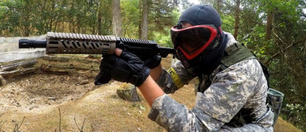 A paintball player wearing protective gear aims a paintball gun in the forest during a game, 2K Desktop Wallpaper