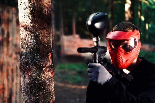 A paintball player wearing a red mask is aiming a paintball gun behind a tree during a game, HD Desktop Wallpaper