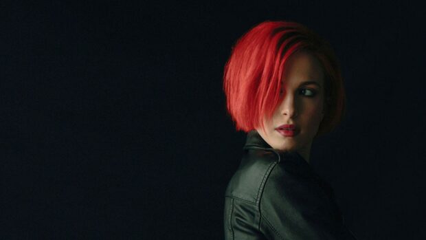 A woman with bright red hair and a leather jacket looks to the side in a dark setting, HD Desktop Wallpaper
