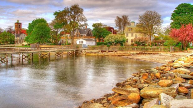 A peaceful village beside a calm river with trees and houses under a cloudy sky, HD Desktop Wallpaper