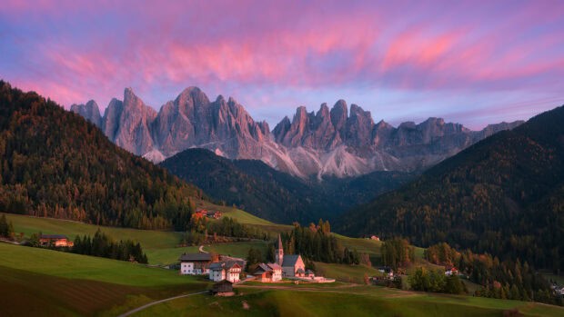 A peaceful village nestled among green hills with mountains in the background during sunset, 2K Desktop Wallpaper