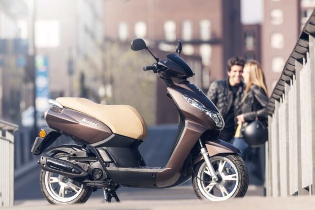 A Peugeot Kisbee scooter is parked on a city street with two people blurred in the background, HD Desktop Wallpaper