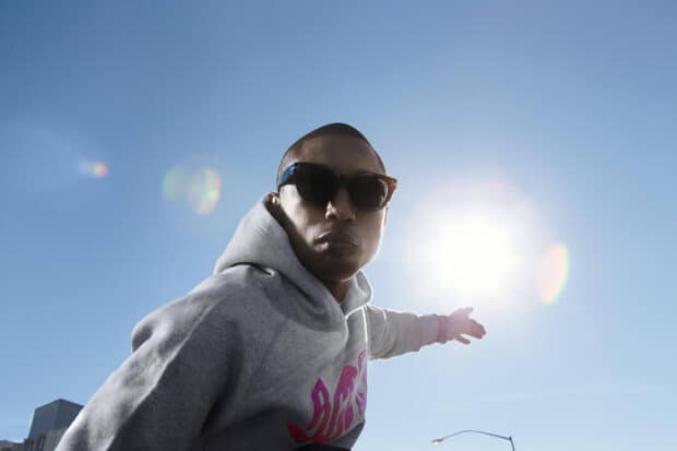 Pharrell Williams is wearing sunglasses and a hoodie while pointing at the bright sun on a clear day, 2K Desktop Wallpaper