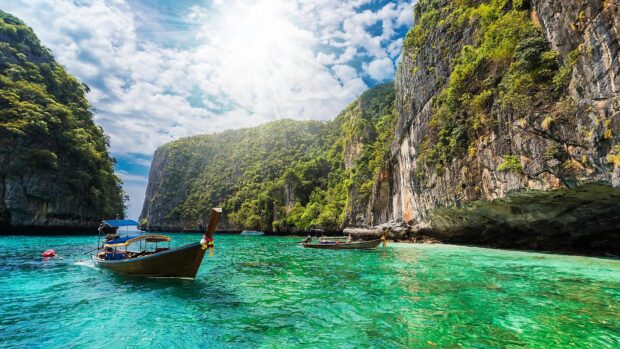 A wooden boat floats on clear turquoise water near limestone cliffs under a bright sky in Phuket, HD Desktop Wallpaper