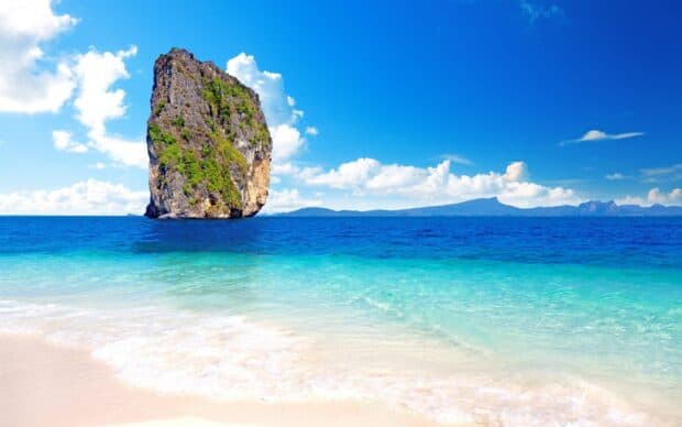 A tall rock formation rises from clear blue waters near a tropical beach in Phuket, 2K Desktop Wallpaper
