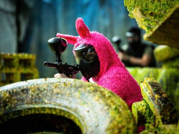 A player in a pink bunny costume is aiming a paintball gun behind cover during a paintball match, HD Desktop Wallpaper