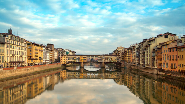 A scenic view of Ponte Vecchio bridge is reflected in the calm Arno river during a cloudy day, 2K Desktop Wallpaper