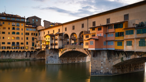 The Ponte Vecchio is captured with warm sunset light reflecting on the water and colorful buildings, 2K Desktop Wallpaper