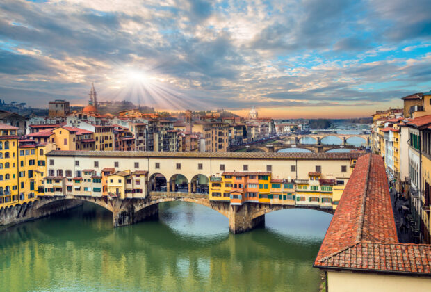 The Ponte Vecchio bridge spans the river under sun rays in a vibrant city, 2K Desktop Wallpaper