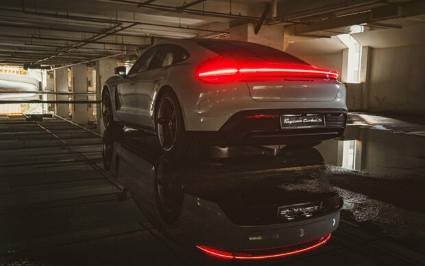A Porsche Taycan with illuminated tail lights is parked in an indoor garage reflecting on a wet floor, 2K Desktop Wallpaper