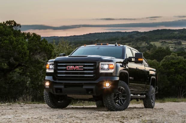 The black GMC Sierra truck is parked on a rugged dirt surface with trees and hills in the background at sunset, HD Desktop Wallpaper