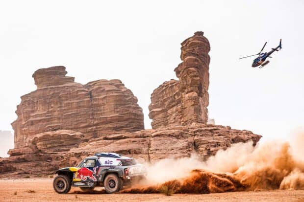 A rally raid vehicle kicks up dust while racing near rocky formations and a helicopter flies above, 2K Desktop Wallpaper