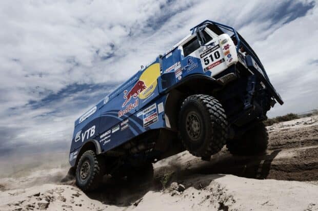 A rally raid truck drives through a sandy desert terrain under a cloudy sky during a race, 2K Desktop Wallpaper