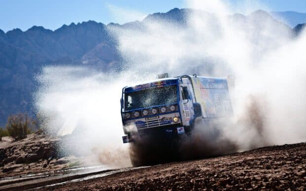 A rally raid truck is speeding through a dusty track with mountains in the background, 2K Desktop Wallpaper