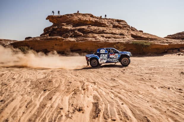 A rally raid car races through a desert landscape with rocky cliffs and photographers, HD Desktop Wallpaper
