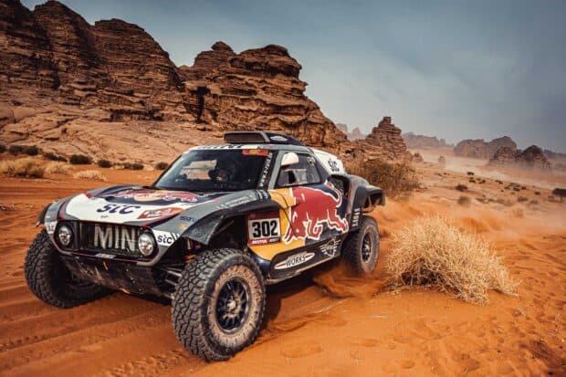 A rally raid vehicle speeds through a desert landscape kicking up sand near rocky formations, HD Desktop Wallpaper