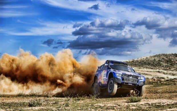 A rally raid vehicle speeds across rough terrain creating a large dust cloud under a blue sky, HD Desktop Wallpaper