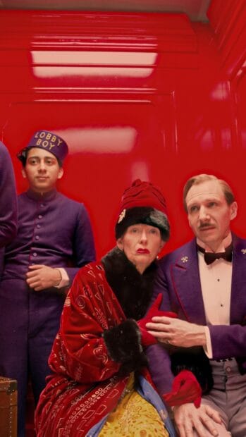 Ralph Fiennes is seen in a purple suit next to two characters dressed in vintage attire in a red room, HD Mobile Wallpaper
