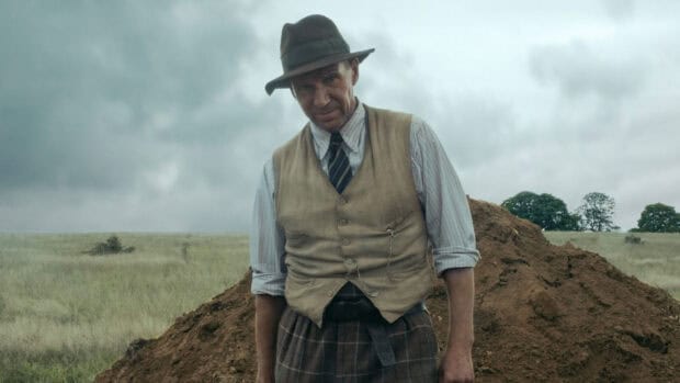 Ralph Fiennes stands outdoors wearing a hat and vest in a grassy field, HD Desktop Wallpaper