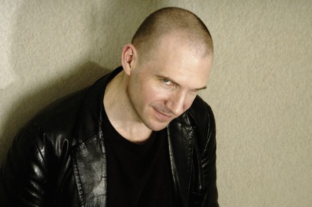 Ralph Fiennes smiles subtly wearing a black leather jacket against a beige wall, 2K Desktop Wallpaper