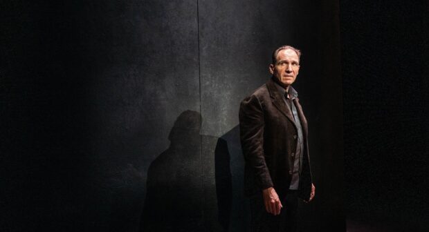 Ralph Fiennes wallpapers shows a man standing against a dark textured wall with a shadow, HD Desktop Wallpaper