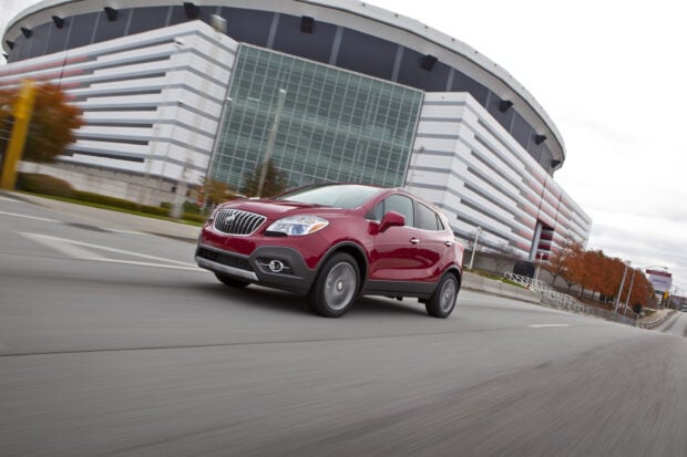 A red Buick Encore is driving past a modern building with autumn trees nearby, 2K Desktop Wallpaper