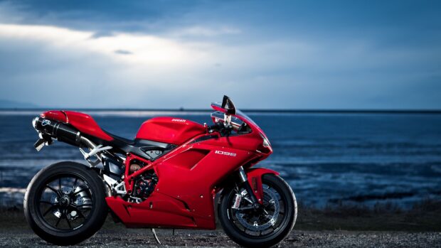 A red Ducati street bike stands near the ocean under a cloudy sky, 4K Desktop Wallpaper