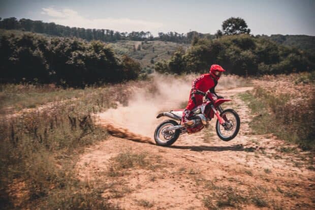 A rider on an enduro motorbike is jumping over a dirt trail kicking up dust in a forest area