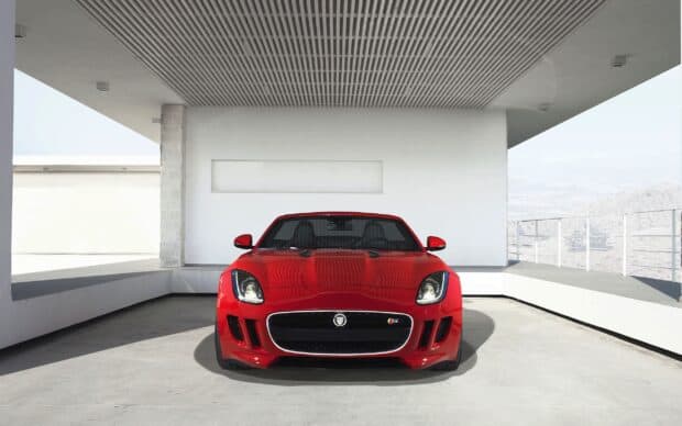 A red Jaguar F Type sports car is parked inside a spacious modern garage with a scenic view outside, 2K Desktop Wallpaper