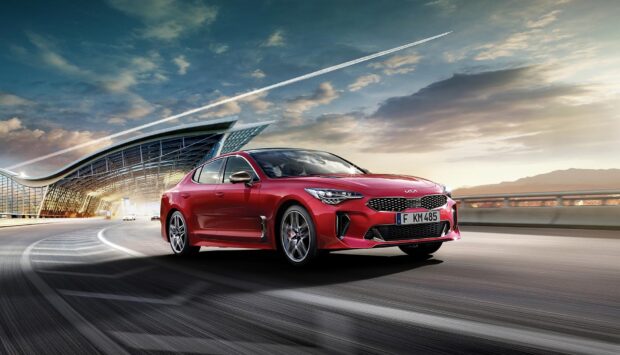 A red Kia Stinger drives past an architectural structure under a partly cloudy sky, HD Desktop Wallpaper